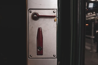 A metal panel featuring a mechanism with a dark red handle and a matching lock component. The panel is mounted on a vertical surface near a subway or train door area, with a blurred background suggesting an urban transit environment.