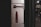 A metal panel featuring a mechanism with a dark red handle and a matching lock component. The panel is mounted on a vertical surface near a subway or train door area, with a blurred background suggesting an urban transit environment.