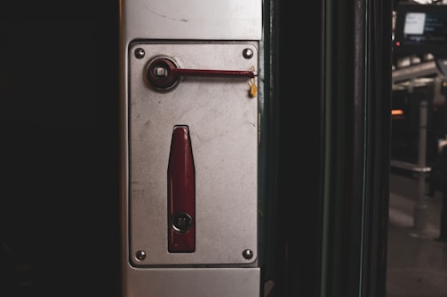 A metal panel featuring a mechanism with a dark red handle and a matching lock component. The panel is mounted on a vertical surface near a subway or train door area, with a blurred background suggesting an urban transit environment.