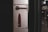 A metal panel featuring a mechanism with a dark red handle and a matching lock component. The panel is mounted on a vertical surface near a subway or train door area, with a blurred background suggesting an urban transit environment.