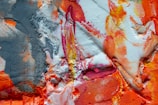 A vibrant abstract painting with a mix of textures and colors, including bold strokes of orange, red, and pink, contrasted with patches of white and shades of gray. The surface appears textured and layered, with some areas appearing smooth while others are rough and uneven.