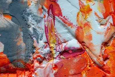 A vibrant abstract painting with a mix of textures and colors, including bold strokes of orange, red, and pink, contrasted with patches of white and shades of gray. The surface appears textured and layered, with some areas appearing smooth while others are rough and uneven.