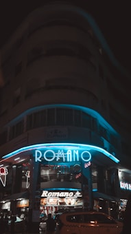 An illuminated storefront with the name 'ROMANO' displayed in bright blue neon lights. The building is multi-storied, and the scene appears to be taken at night. The store seems to be a restaurant or an ice cream parlor, as suggested by the ice cream cone signage and imagery. A vehicle is parked in front of the building.