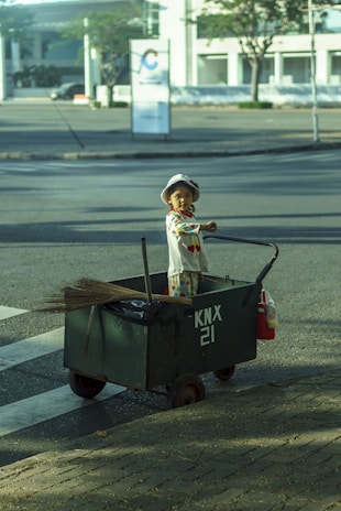 A young child wearing a light-colored hat and patterned clothes stands inside a large green cart on an empty street. The cart has 'KNX 21' written on its side and contains a broomstick and some cleaning tools. The background shows a quiet urban area with some buildings and trees.