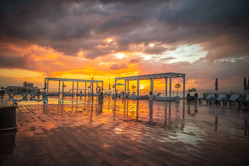 A rooftop lounge area overlooks a vivid sunset. The sky is filled with vibrant orange and yellow hues, contrasting with dark, dramatic clouds. Reflections glisten on the wet wooden floor, while several lounge chairs and white pergolas are distributed across the deck. Potted plants add a touch of greenery to the modern, serene setting.