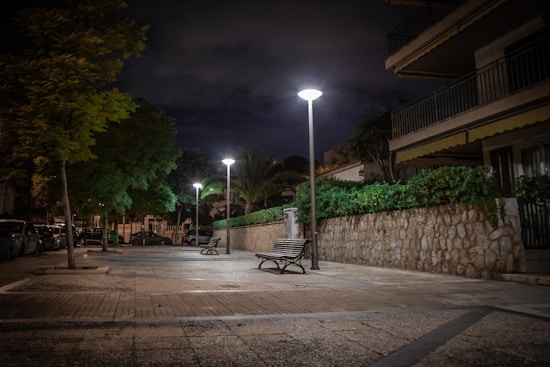 A quiet urban street scene at night with evenly spaced streetlights casting pools of light on the pavement. Trees line one side of the street, and a building with balconies is on the other. Benches are placed along the walkway, offering a place to sit. Cars are parked along the street under the glow of the streetlights.