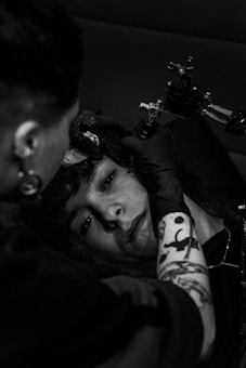 A person is getting a tattoo on their forehead by a tattoo artist. The image is in black and white, adding a dramatic effect. The artist is holding a tattoo machine, and the client appears calm and focused. The tattoo artist has visible tattoos on their arm and is wearing gloves.