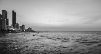 A monochrome photograph featuring a coastal cityscape during evening or twilight. High-rise buildings and city lights line the left side of the frame. The ocean dominates the right side, with gentle waves. A few people can be seen in the shallow waters near the shore.