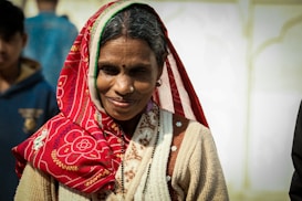 An elderly woman with a warm smile wears a vibrant red patterned headscarf and a cream-colored sweater. She has a nose pin and a bindi on her forehead, and her hair is visible under the scarf. The background includes some blurred figures, suggesting an outdoor setting.