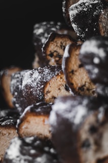 Several baked pastries with a dark, crusty exterior dusted with powdered sugar. The pastries reveal a lighter, crumbly interior with visible dark chocolate chips or dried fruit pieces. The focus is shallow, emphasizing the textures and details of the pastries against a dark background.