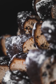 Several baked pastries with a dark, crusty exterior dusted with powdered sugar. The pastries reveal a lighter, crumbly interior with visible dark chocolate chips or dried fruit pieces. The focus is shallow, emphasizing the textures and details of the pastries against a dark background.