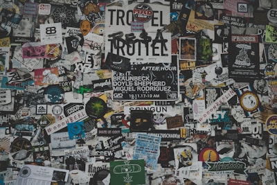 A chaotic collage of various stickers, posters, and flyers layered on a surface. The overlapping images and texts create a dense and vibrant atmosphere, featuring a mix of artistic designs, advertisements, and messages in multiple languages. Some elements include band names, events, artistic logos, and social tags.