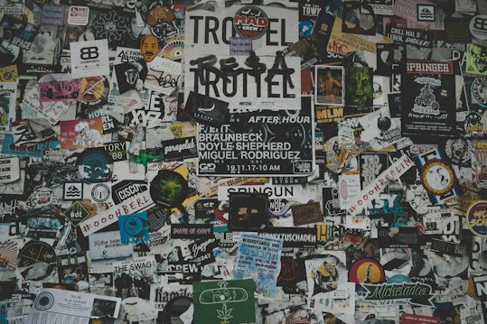 A chaotic collage of various stickers, posters, and flyers layered on a surface. The overlapping images and texts create a dense and vibrant atmosphere, featuring a mix of artistic designs, advertisements, and messages in multiple languages. Some elements include band names, events, artistic logos, and social tags.