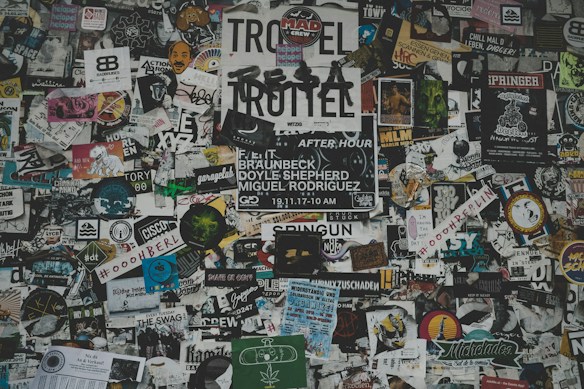 A chaotic collage of various stickers, posters, and flyers layered on a surface. The overlapping images and texts create a dense and vibrant atmosphere, featuring a mix of artistic designs, advertisements, and messages in multiple languages. Some elements include band names, events, artistic logos, and social tags.