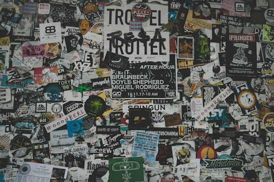 A chaotic collage of various stickers, posters, and flyers layered on a surface. The overlapping images and texts create a dense and vibrant atmosphere, featuring a mix of artistic designs, advertisements, and messages in multiple languages. Some elements include band names, events, artistic logos, and social tags.