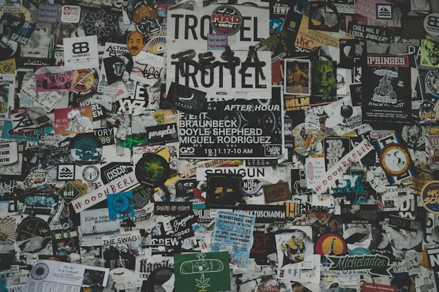 A chaotic collage of various stickers, posters, and flyers layered on a surface. The overlapping images and texts create a dense and vibrant atmosphere, featuring a mix of artistic designs, advertisements, and messages in multiple languages. Some elements include band names, events, artistic logos, and social tags.