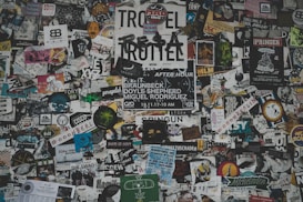 A chaotic collage of various stickers, posters, and flyers layered on a surface. The overlapping images and texts create a dense and vibrant atmosphere, featuring a mix of artistic designs, advertisements, and messages in multiple languages. Some elements include band names, events, artistic logos, and social tags.