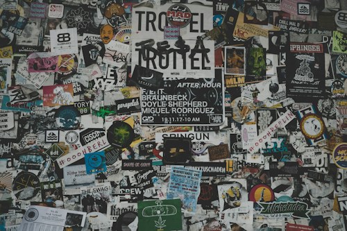 A chaotic collage of various stickers, posters, and flyers layered on a surface. The overlapping images and texts create a dense and vibrant atmosphere, featuring a mix of artistic designs, advertisements, and messages in multiple languages. Some elements include band names, events, artistic logos, and social tags.