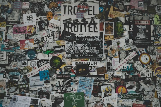 A chaotic collage of various stickers, posters, and flyers layered on a surface. The overlapping images and texts create a dense and vibrant atmosphere, featuring a mix of artistic designs, advertisements, and messages in multiple languages. Some elements include band names, events, artistic logos, and social tags.