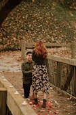 A couple is on a wooden bridge covered with rose petals and surrounded by fallen leaves. One person is kneeling, holding the hand of the other, who is standing. Small candles line the side of the bridge, creating a romantic atmosphere.