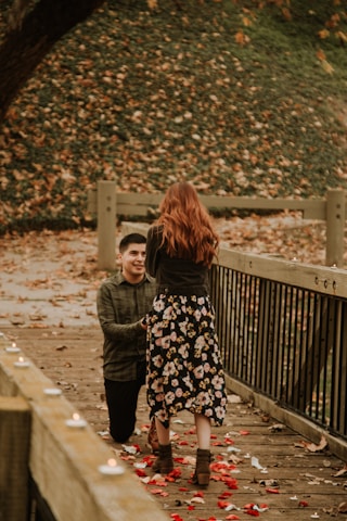 A couple is on a wooden bridge covered with rose petals and surrounded by fallen leaves. One person is kneeling, holding the hand of the other, who is standing. Small candles line the side of the bridge, creating a romantic atmosphere.