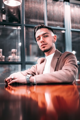 A young man with styled hair and facial hair is seated at a polished wooden table. He is wearing a brown blazer over a white shirt and accessorized with a watch and rings. The background consists of a modern, industrial-style setting with grid-like panels and ambient lighting.