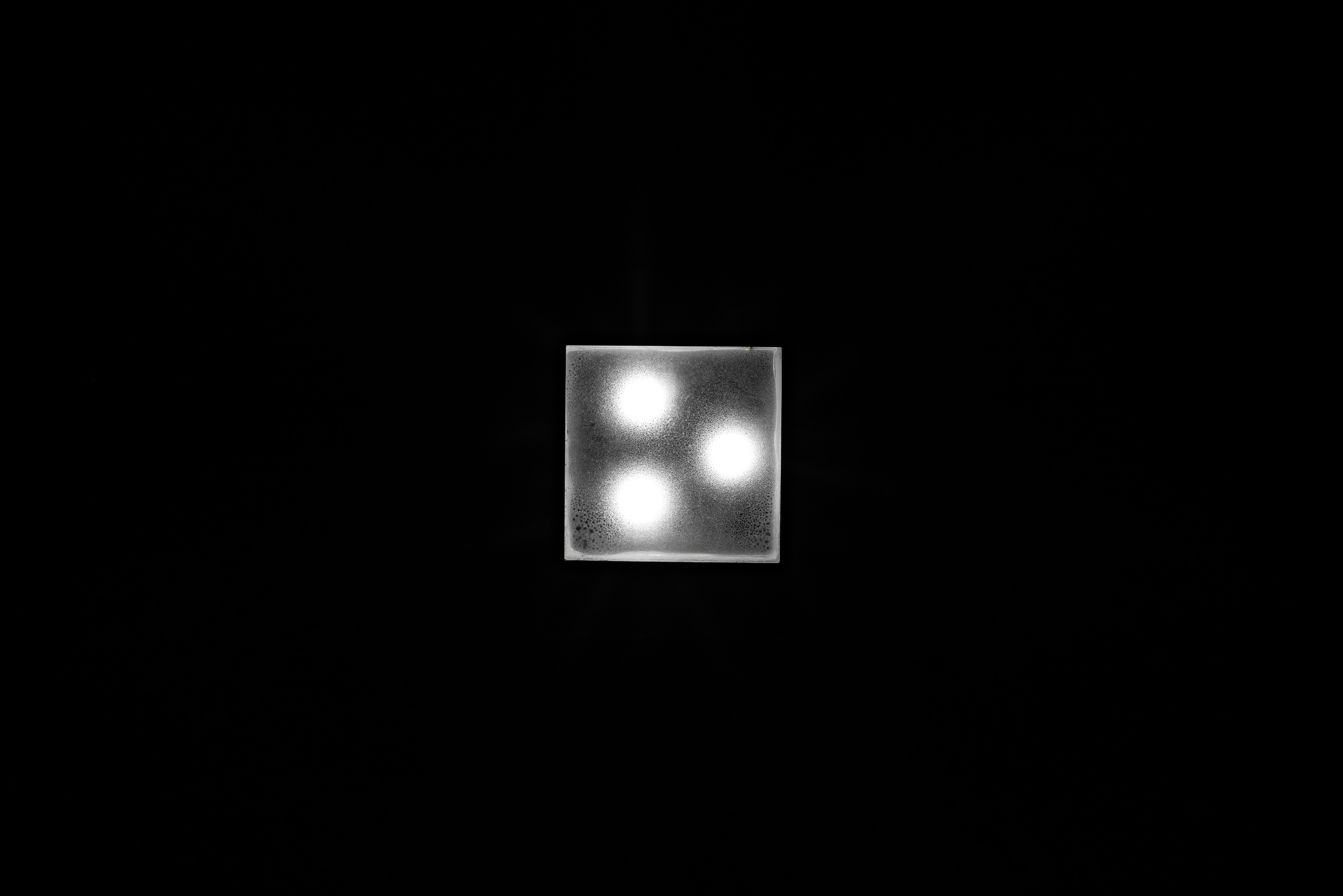 A square light fixture with three glowing bulbs set in a triangular arrangement against a dark background. The fixture is brightly illuminated, creating a sharp contrast with the surrounding darkness.