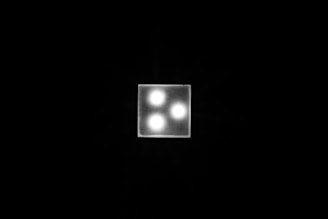 A square light fixture with three glowing bulbs set in a triangular arrangement against a dark background. The fixture is brightly illuminated, creating a sharp contrast with the surrounding darkness.