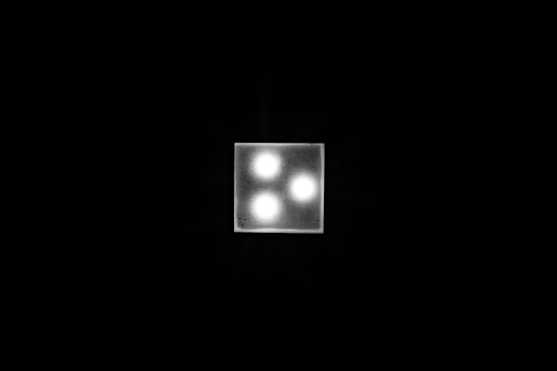 A square light fixture with three glowing bulbs set in a triangular arrangement against a dark background. The fixture is brightly illuminated, creating a sharp contrast with the surrounding darkness.