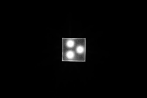 A square light fixture with three glowing bulbs set in a triangular arrangement against a dark background. The fixture is brightly illuminated, creating a sharp contrast with the surrounding darkness.
