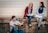 Three young people are sitting and smiling with open books in their laps, enjoying a moment together. They are sitting on a wooden ledge against a light-colored corrugated metal wall. The mood appears relaxed and joyful, with a sense of camaraderie.