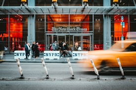 A bustling urban scene outside the entrance of The New York Times building, featuring people walking past NYPD barriers. A yellow taxi speeds by in the foreground, creating a motion blur effect. The building has a modern glass facade with large windows, and warm lighting inside contrasts with the cool exterior.