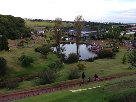 A lush green park landscape features a small lake surrounded by people. Trees and shrubbery are scattered throughout the area, with pathways for walking. A crowd gathers near the lake, and a large gazebo is visible in the background.