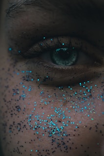 A close-up of a human eye, surrounded by small, scattered specks of blue and black glitter on the skin. The skin texture and details of the eye, including the eyelashes, are visible.