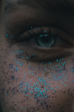 A close-up of a human eye, surrounded by small, scattered specks of blue and black glitter on the skin. The skin texture and details of the eye, including the eyelashes, are visible.