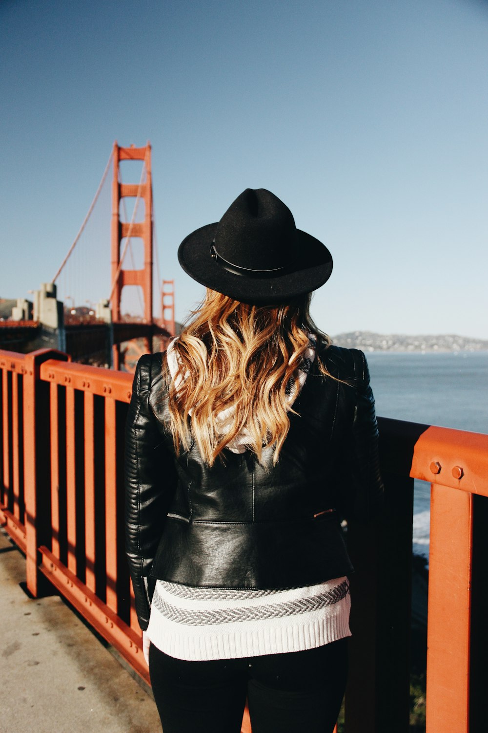 woman wearing black leather jacket and hat while facing red bridge during daytime