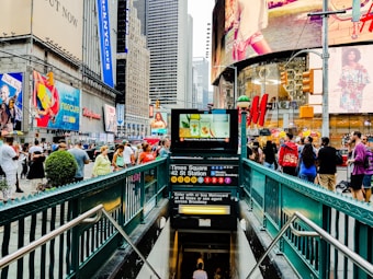A bustling urban scene in Times Square featuring a subway entrance with signs for various train lines. Large digital billboards and advertisements cover the surrounding buildings, and a crowd of people is visible, showcasing a vibrant, busy atmosphere.