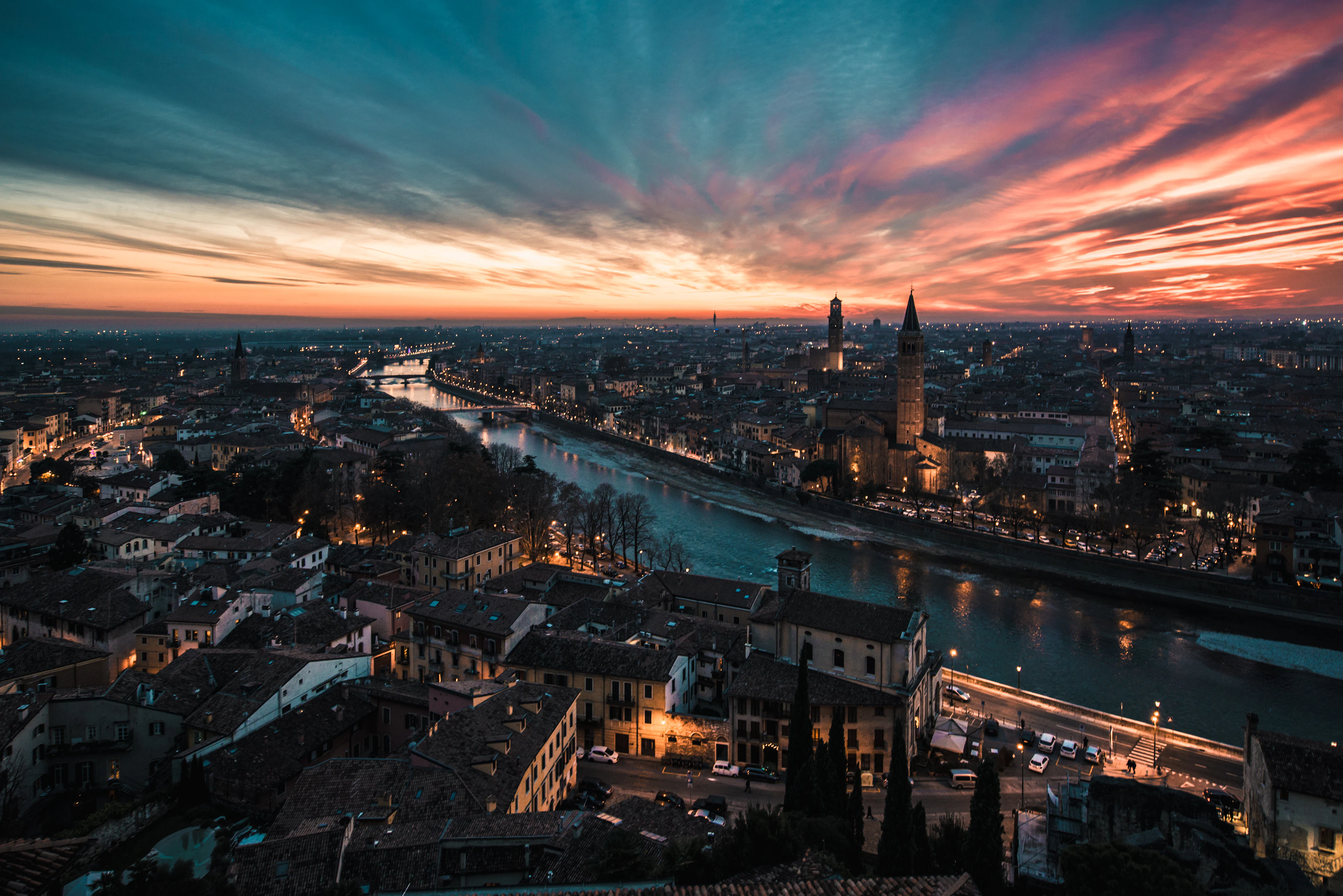 Dusk panorama of Verona from Castel San Pietro, the Adige river curving through the terracotta rooftops as the city lights come on
