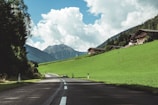 A country road stretches through a picturesque rural landscape, flanked by lush green fields and dense forests. In the background, mountains rise under a blue sky dotted with fluffy white clouds. Clustered on a hillside, charming wooden houses add a rustic touch to the scene.