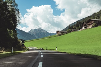A country road stretches through a picturesque rural landscape, flanked by lush green fields and dense forests. In the background, mountains rise under a blue sky dotted with fluffy white clouds. Clustered on a hillside, charming wooden houses add a rustic touch to the scene.