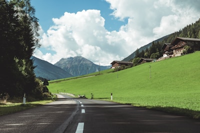 A country road stretches through a picturesque rural landscape, flanked by lush green fields and dense forests. In the background, mountains rise under a blue sky dotted with fluffy white clouds. Clustered on a hillside, charming wooden houses add a rustic touch to the scene.