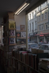 A cozy bookstore interior with shelves filled with books, some of which are stacked on top. The store has large windows showing the street outside, capturing the exterior of nearby buildings.
