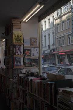A cozy bookstore interior with shelves filled with books, some of which are stacked on top. The store has large windows showing the street outside, capturing the exterior of nearby buildings.