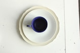 A dark blue ceramic cup is centrally placed on a white, fluted plate, which itself is atop a scalloped beige plate. The arrangement sits on a white table surface with some minor black specks and imperfections.