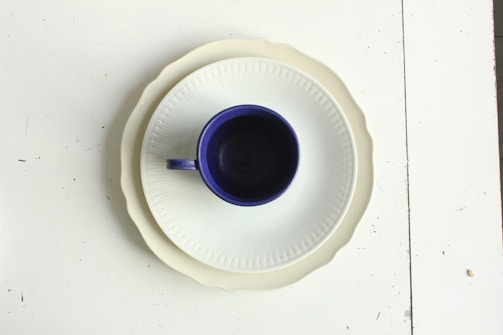 A dark blue ceramic cup is centrally placed on a white, fluted plate, which itself is atop a scalloped beige plate. The arrangement sits on a white table surface with some minor black specks and imperfections.