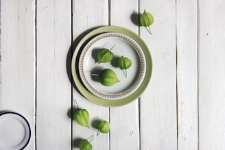 A rustic wooden surface with a set of plates in the center, featuring a decorative green outer plate and a smaller patterned white plate on top. Several green, husk-covered fruits, possibly tomatillos, are artfully arranged both on the plates and scattered around them.
