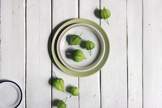 A rustic wooden surface with a set of plates in the center, featuring a decorative green outer plate and a smaller patterned white plate on top. Several green, husk-covered fruits, possibly tomatillos, are artfully arranged both on the plates and scattered around them.