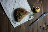 A rustic loaf of brown bread rests on crumpled white parchment paper with sprigs of fresh herbs scattered around. A small jar filled with what appears to be butter sits nearby, complemented by a knife placed on the wooden table surface.