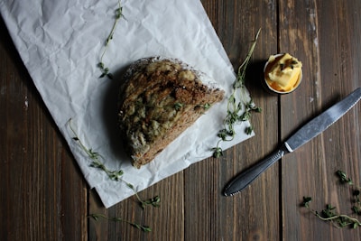 A rustic loaf of brown bread rests on crumpled white parchment paper with sprigs of fresh herbs scattered around. A small jar filled with what appears to be butter sits nearby, complemented by a knife placed on the wooden table surface.
