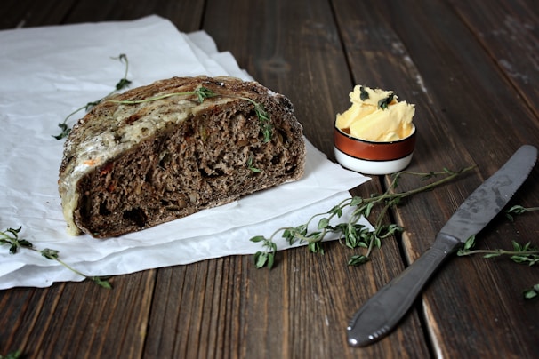 A rustic piece of brown bread is partially covered with melted cheese and garnished with fresh herbs. It rests on white parchment paper. Beside it, there's a small round bowl filled with butter topped with herbs. A knife with a metallic handle is placed nearby on a wooden surface, surrounded by sprigs of herbs.